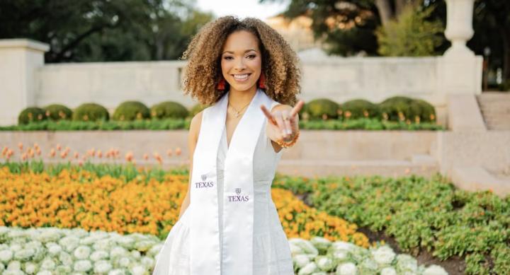 Chanel Williams wears a white convocation sash and makes a Hook 'em Horns hand sign in front of a flowerbed on the UT Austin campus.